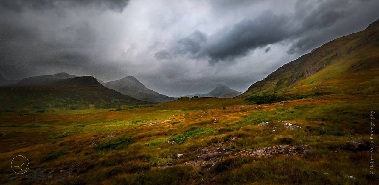 A wide green valley between low mountains, rain clouds hanging over the tops
