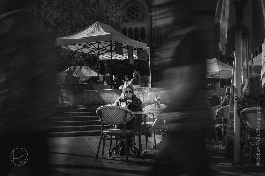 A café terrace under umbrellas, a woman sitting still while the crowd blurs past