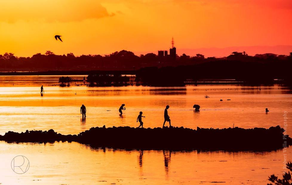An orange sunset over shallow water, people wading, birds crossing the sky