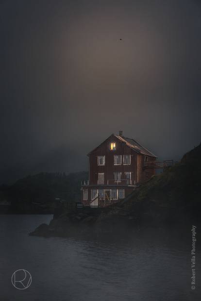 A red timber house on a rocky shore, fog and a low moon behind it